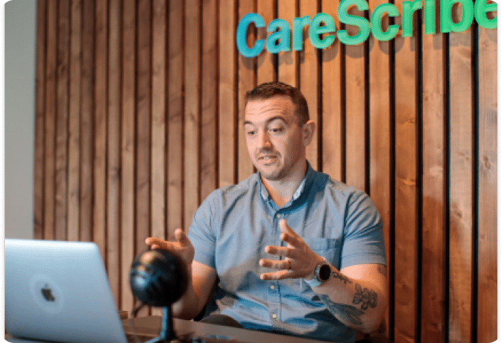 A photo of Stefano showing a demo during a TalkType webinar, sitting in front of a CareScribe sign on a wooden wall.